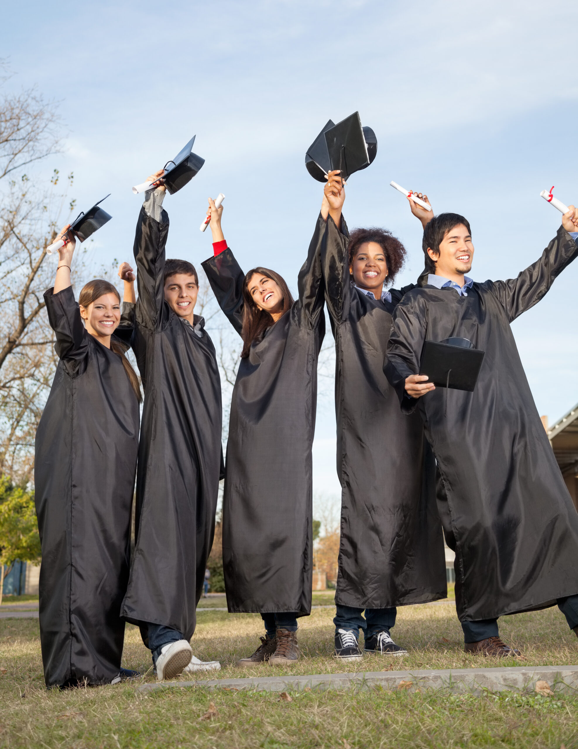 Full length of happy students with diplomas celebrating success on graduation day at college campus